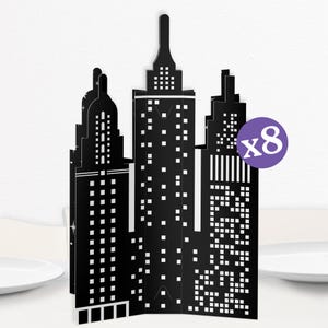 May include: Black and white paper cutouts of city skyscrapers. The buildings have windows and are arranged in a row. A purple circle with the number "x8" is in the upper right corner. The cutouts are on a white surface.