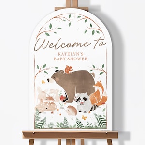 May include: A white, arched sign on an easel with the words "Welcome to Katelyn's Baby Shower" in brown script. The sign features illustrations of woodland animals, including a bear, fox, deer, and raccoon, with green foliage accents.
