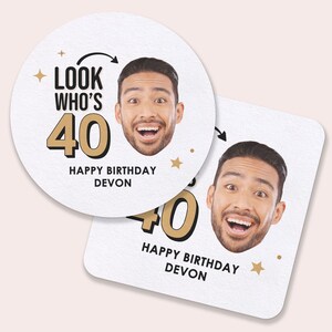 May include: White round and square coasters with a photo of a smiling person. The coasters have the text "LOOK WHO'S 40" in black and gold, with "HAPPY BIRTHDAY DEVON" below. Gold stars and arrows are also included.