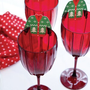 Puede incluir: Tres copas de vino rojas con un dise&ntilde;o acanalado, cada una adornada con un adorno de &aacute;rbol de Navidad en forma de su&eacute;ter verde. Una servilleta de lunares rojos y blancos est&aacute; en el fondo. Decoraci&oacute;n festiva navide&ntilde;a.