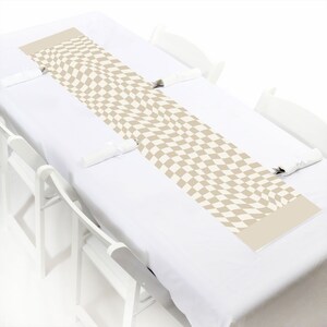 May include: A beige and white checkered table runner is centered on a white tablecloth. The runner has a wavy pattern and is rectangular. White chairs are positioned around the table, with rolled napkins and silverware.