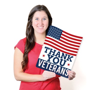Puede incluir: Una mujer sostiene un cartel con una bandera estadounidense y el texto "Thank You Veterans" en letras azules y rojas.