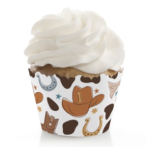 May include: A cupcake with white frosting in a decorative cowboy-themed wrapper. The wrapper features a brown cowboy hat with the number 1, horseshoes, stars, and cowhide spots. The cupcake is on a white background.