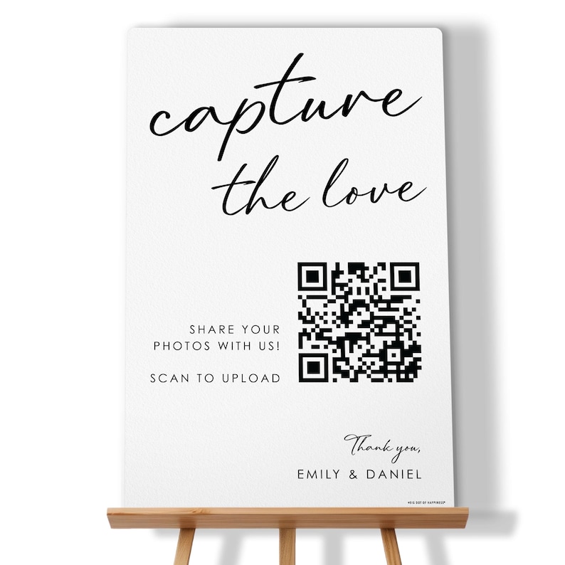 May include: A white rectangular sign with black text that reads capture the love, SHARE YOUR PHOTOS WITH US! SCAN TO UPLOAD, and Thank you, EMILY & DANIEL. The sign features a large black and white QR code in the center and rests on a wooden easel.