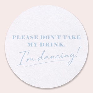 May include: A round white coaster with the text "PLEASE DON'T TAKE MY DRINK, I'm dancing!" in light blue. The coaster has a textured surface and is suitable for placing drinks on.
