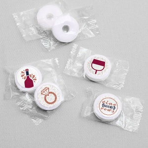 May include: Five round white mints individually wrapped in clear plastic. Three mints feature printed designs: a red wine bottle, a diamond engagement ring, a wine glass, and the text "vino BEFORE vows" in rose gold and black lettering.