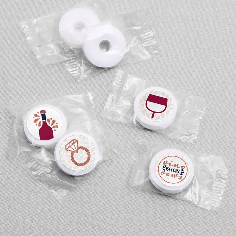 May include: Five round white mints individually wrapped in clear plastic. Three mints feature printed designs: a red wine bottle, a diamond engagement ring, a wine glass, and the text "vino BEFORE vows" in rose gold and black lettering.