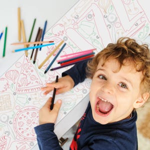 Puede incluir: Un ni&ntilde;o con pelo rizado sonr&iacute;e ampliamente, sosteniendo un l&aacute;piz negro sobre una hoja para colorear con ilustraciones de pizza y comida. Varios l&aacute;pices de colores est&aacute;n esparcidos sobre la mesa.