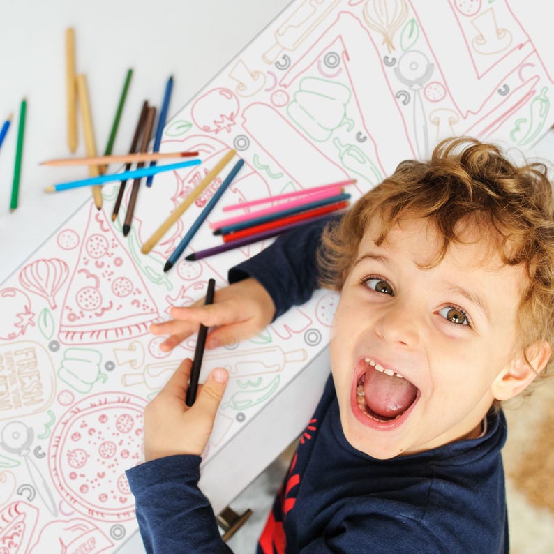 Puede incluir: Un ni&ntilde;o con pelo rizado sonr&iacute;e ampliamente, sosteniendo un l&aacute;piz negro sobre una hoja para colorear con ilustraciones de pizza y comida. Varios l&aacute;pices de colores est&aacute;n esparcidos sobre la mesa.