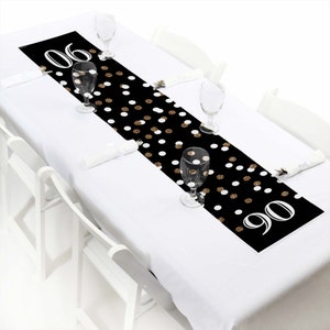 May include: Black and white table runner with gold glitter polka dots and the numbers "90" printed on the end. The table runner is on a white table with white chairs.