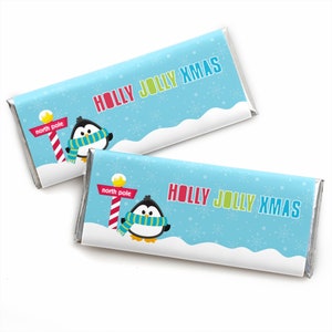 May include: Two rectangular chocolate bars wrapped in light blue paper featuring a white snowy landscape, snowflakes, a penguin wearing a teal and yellow striped scarf, a red and white North Pole sign, and the text HOLLY JOLLY XMAS.