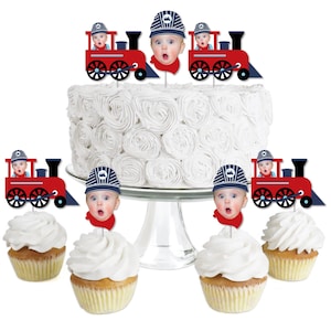May include: Red and blue train cake toppers with a baby's face on each. The toppers are on a white cake and cupcakes.