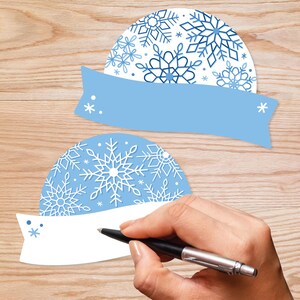 May include: Two blue and white snowflake-patterned paper ornaments with blank white banners attached. The banners are ready for writing with a pen.