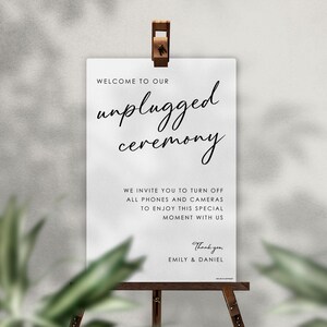 May include: A white sign with the words "unplugged ceremony" in elegant script. The sign is held by a wooden easel and includes a message asking guests to turn off phones and cameras. The sign is for a wedding or special event.