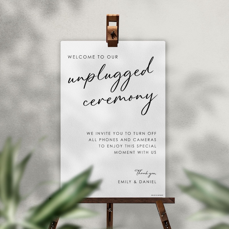 May include: A white sign with the words "unplugged ceremony" in elegant script. The sign is held by a wooden easel and includes a message asking guests to turn off phones and cameras. The sign is for a wedding or special event.