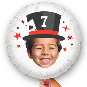 May include: A white, round foil balloon featuring a smiling child's face. The balloon has a black top hat with a red band and the number 7. An ace of spades card and red starbursts decorate the balloon.