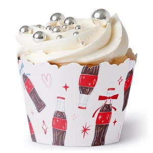 May include: A cupcake with white frosting and silver ball sprinkles. The cupcake liner is white with a repeating pattern of cola bottles in red and brown, along with hearts and stars.