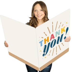May include: A large, open greeting card with the words "THANK YOU!" in colorful, stylized lettering. The card has a cream-colored background with a brown border and is held open, showcasing the message.