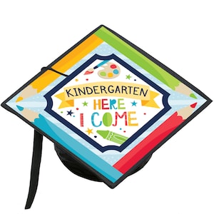 Puede incluir: Un gorro de graduación negro con un diseño colorido que incluye lápices de colores, pinceles y el texto "Kindergarten Here I Come".