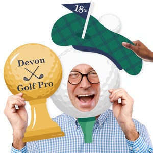 May include: A golf-themed photo booth prop with a white golf ball with a hole for a face, a green and blue golf hat, and a gold trophy with the text 