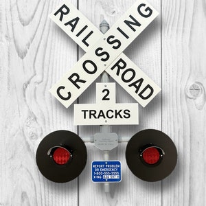 May include: A white railroad crossing sign with black lettering that reads "RAIL CROSSING ROAD" and "2 TRACKS". Two red flashing lights are attached to the sign. A blue sign below the lights reads "REPORT PROBLEM OR EMERGENCY 1-800-555-5555 X-ING 836 597 H".