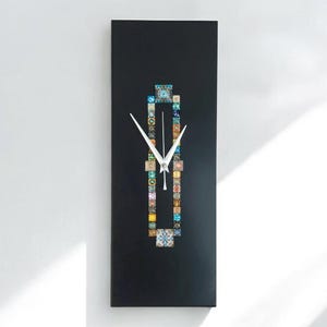 May include: A rectangular black wall clock with white hands. The clock face has a colourful, geometric design. The clock is mounted on a white wall, with shadows from sunlight.