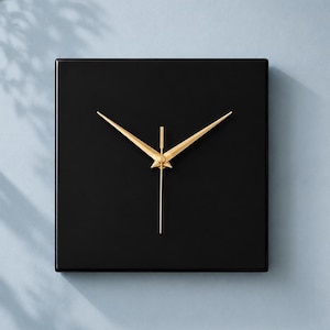 Könnte beinhalten: Eine quadratische, schwarze Wanduhr mit goldfarbenen Zeigern. Die Uhr hat ein minimalistisches Design und ist an einer hellblauen Wand befestigt. Die Zeiger der Uhr sind dünn und elegant und sorgen für eine moderne Ästhetik. Maße in Zentimetern.