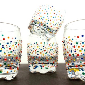Op de afbeelding: Vier heldere drinkglazen met kleurrijke stippen in rood, geel, blauw en groen. De glazen zijn in een groep gerangschikt, met &eacute;&eacute;n glas bovenop twee andere. De glazen staan op een donkere ondergrond.