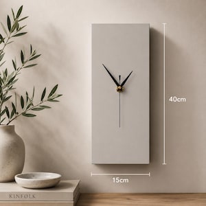 May include: A rectangular, light gray wall clock with black hands and a gold-colored center. The clock is 15cm wide and 40cm tall. The clock is mounted on a light beige wall. A vase with greenery is to the left of the clock.