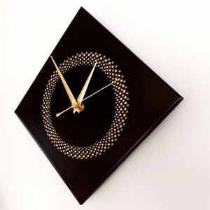 May include: A square black wall clock with gold hands and a gold and silver dot design forming a circle. The clock is mounted on a white wall. The clock's hands show the time.