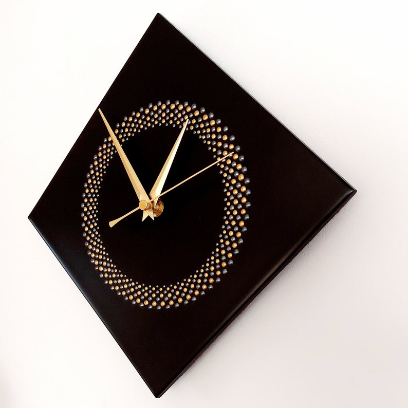 May include: A square black wall clock with gold hands and a gold and silver dot design forming a circle. The clock is mounted on a white wall. The clock's hands show the time.