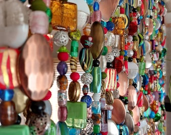 May include: A colorful beaded curtain with a variety of shapes and sizes. The beads are in shades of red, green, blue, gold, and copper. Some beads are round, others are square or oval. The curtain is hanging in front of a window.