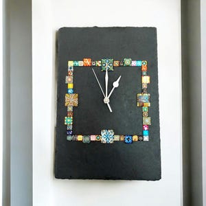 May include: A rectangular black slate clock with a colourful mosaic border. The mosaic is composed of various square tiles in shades of blue, green, yellow, and pink. The clock hands are white, and the clock is mounted on a white wall.