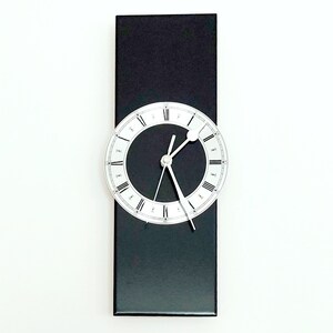 Peut inclure: Une horloge murale moderne avec une base rectangulaire noire et un cadran circulaire. Le cadran est noir avec des aiguilles argentées et des chiffres romains. L'horloge est fixée sur un mur blanc.