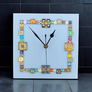 May include: Square wall clock with a light blue face and black hands. The clock is framed with colourful square tiles. The tiles have various patterns and colours, including orange, yellow, blue, and green. The clock is a decorative item.