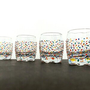 Op de afbeelding: Vier heldere, tonvormige drinkglazen met kleurrijke stippen. De glazen zijn in een rij gerangschikt, elk iets groter dan de vorige. De stippen zijn in verschillende kleuren, waaronder rood, blauw, geel en groen.