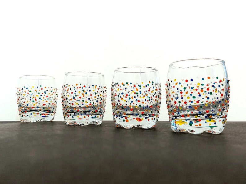 Op de afbeelding: Vier heldere, tonvormige drinkglazen met kleurrijke stippen. De glazen zijn in een rij gerangschikt, elk iets groter dan de vorige. De stippen zijn in verschillende kleuren, waaronder rood, blauw, geel en groen.