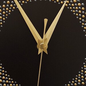 May include: Close-up of a black clock face with gold and silver beaded accents forming a circle. The clock hands are gold. The design is minimalist and elegant.