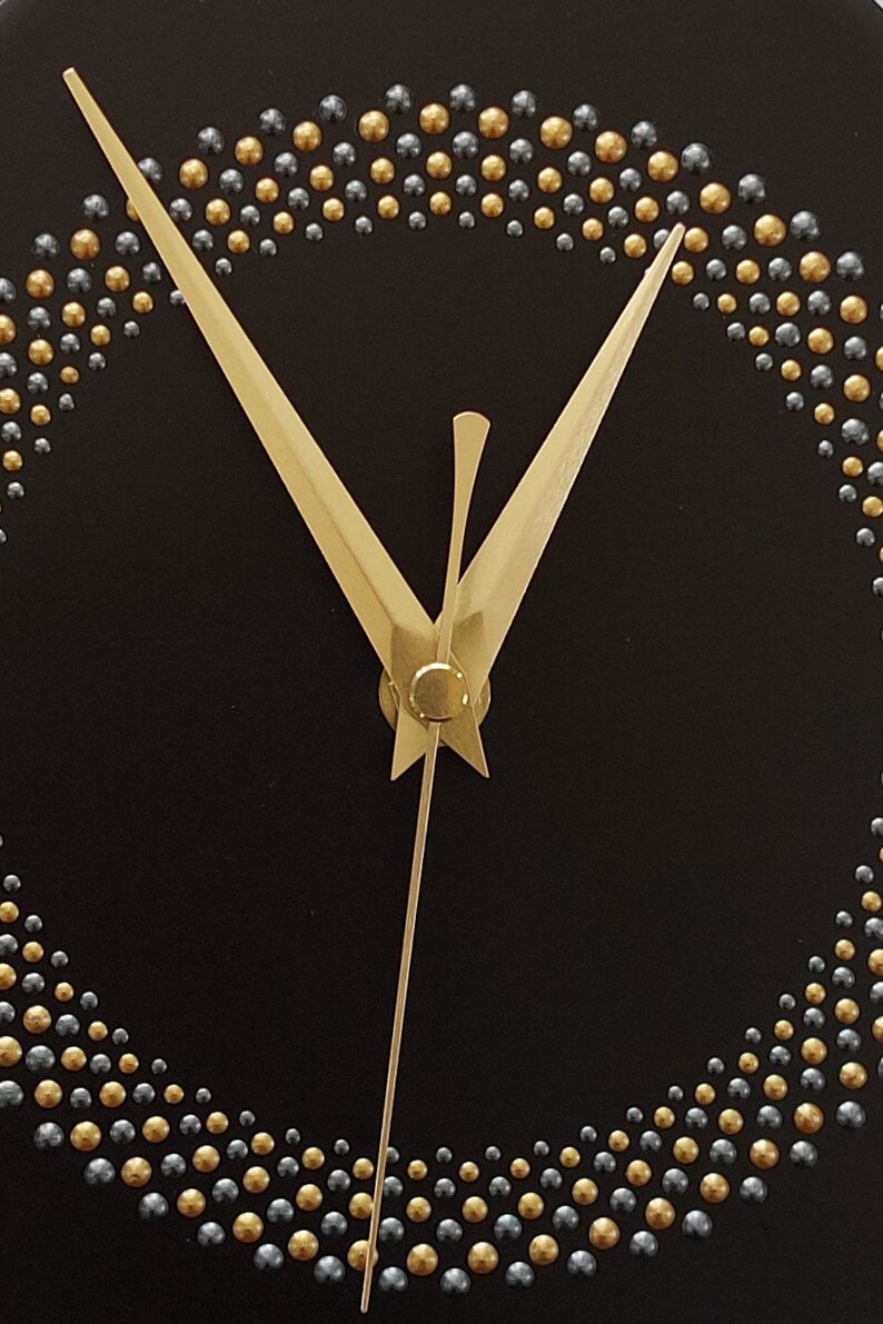 May include: Close-up of a black clock face with gold and silver beaded accents forming a circle. The clock hands are gold. The design is minimalist and elegant.