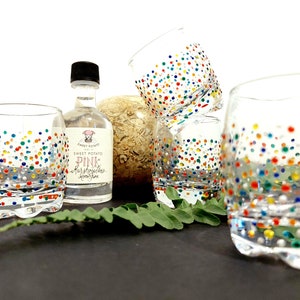Op de afbeelding: Transparante drinkglazen versierd met kleurrijke stippen. In het midden staat een kleine fles Sweet Potato Pink Marshmallows Moonshine. De glazen zijn gerangschikt met een varen en een decoratieve vaas.