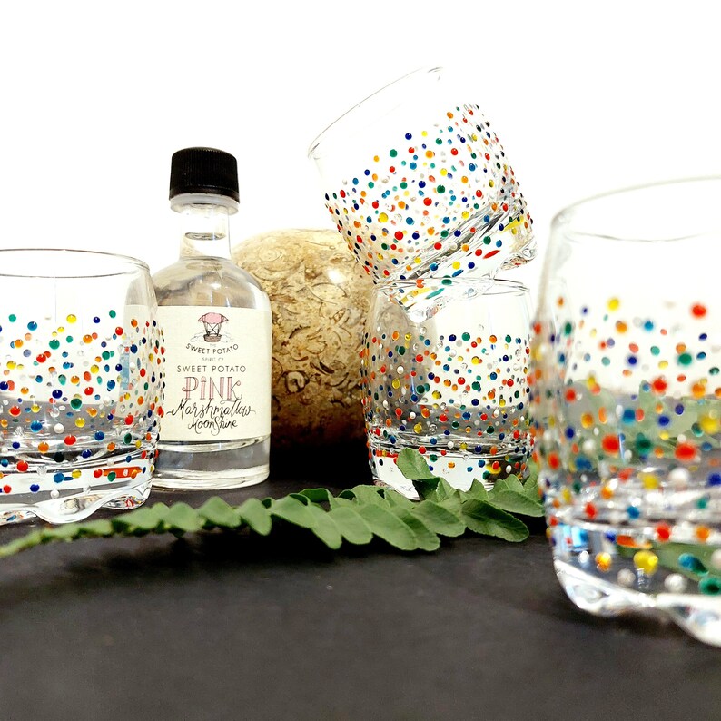 Op de afbeelding: Transparante drinkglazen versierd met kleurrijke stippen. In het midden staat een kleine fles Sweet Potato Pink Marshmallows Moonshine. De glazen zijn gerangschikt met een varen en een decoratieve vaas.