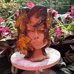 May include: A ceramic planter shaped like a human face, featuring textured, speckled clay skin and adorned with sculpted, glossy autumn leaves in shades of orange, yellow, and deep brown that frame the face and cascade down the sides.