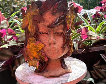 May include: A ceramic planter shaped like a human face, featuring textured, speckled clay skin and adorned with sculpted, glossy autumn leaves in shades of orange, yellow, and deep brown that frame the face and cascade down the sides.
