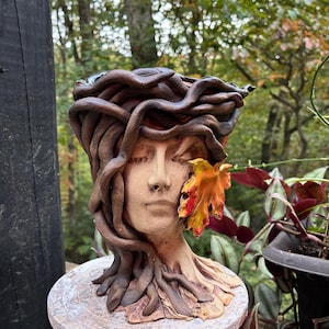 May include: A ceramic planter shaped like a human head, adorned with sculpted brown snakes winding around the top and a single, vibrant orange and yellow autumn leaf attached to the side of the face, featuring closed eyes and a neutral expression.