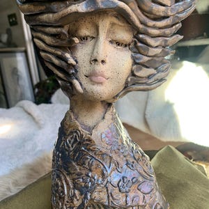 May include: A ceramic sculpture of a woman's bust with flowing hair and a detailed floral patterned collar. The sculpture is primarily brown with speckled details and a matte finish, showcasing an artistic and handcrafted aesthetic.
