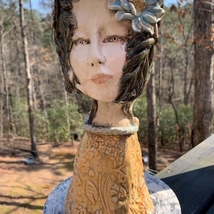 May include: A ceramic sculpture of a woman's face with a floral design. The face is painted with neutral tones, with a blue flower on the head and green leaves. The base is a textured, yellow-orange color.