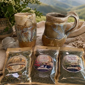 May include: Two handcrafted ceramic mugs with earthy brown and blue glazes, a metal tea infuser, and three clear pouches of loose-leaf tea labeled Garden Summer Solstice, Catch Your Breath, and Settle the Belly, arranged on a textured fabric.