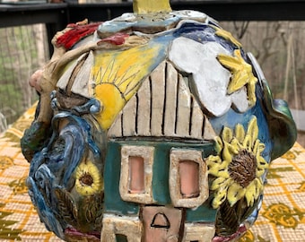 May include: A spherical ceramic sculpture featuring a relief design of a house with a teal exterior, a pink door, and two windows. The surface includes a yellow star at the top, a sunflower, a sailboat on blue waves, and white clouds against a blue sky.