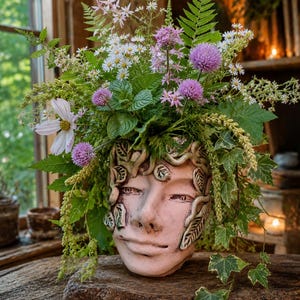 May include: A ceramic planter shaped like a human face with sculpted leaves and vines, filled with a lush arrangement of purple chive blossoms, white daisies, green ferns, and trailing ivy leaves, creating a vibrant and natural botanical display.
