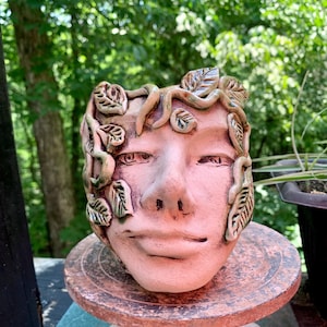 May include: A ceramic planter shaped like a human face with sculpted vines and leaves wrapping around the forehead and sides. The piece features a light pinkish-tan finish with subtle green glazing on the foliage and a neutral facial expression.